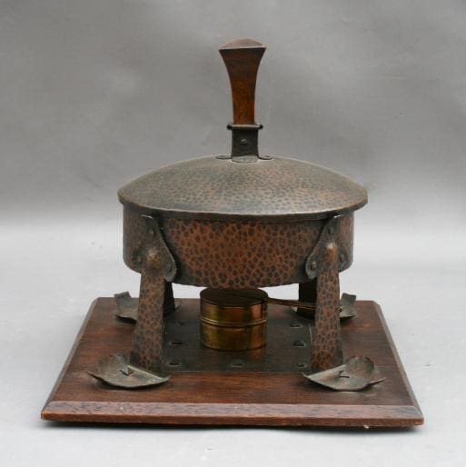Antique metal Arts And Crafts Chafing Dish on a wooden base, featuring embossed designs and a central handle on the lid.
