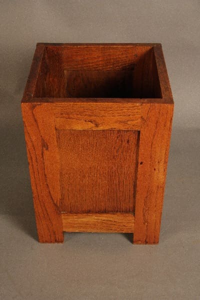 A 11885. Arts And Crafts Waste Basket with a visible grain pattern and a simple panel design on the side, shown against a gray background.