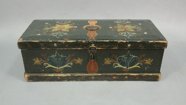 An antique black wooden trunk decorated with floral patterns in gold, featuring a central metal handle and a hanging lock.