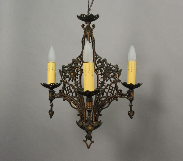 An 1920's Spanish Revival chandelier with lion motif, featuring three arms each holding a candle-shaped light bulb, suspended against a gray background.