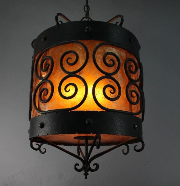 14077. 1 Of 2 Antique Wrought Iron Drum with Replaced Mica chandelier with swirling patterns illuminated from within, casting a warm glow against a gray background.