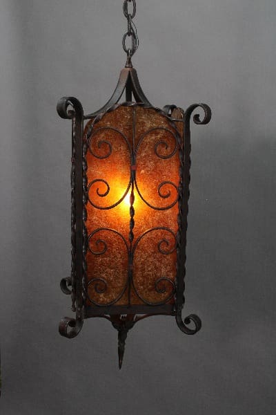 A lit Spanish Revival 1920's pendant with mica detailing and an amber glass shade, suspended by a chain against a gray background.