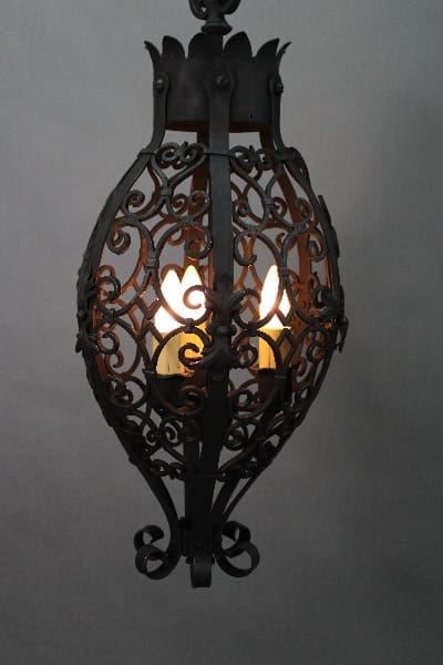 14254. 1920's Wrought Iron Pendant With Three Lights with a delicate lattice design, emitting a warm glow from its central light against a gray background.
