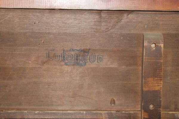 Faded blue stamp on a 14329. Signed Coronado Sideboard with brown and rusty tones, showing worn text and a graphical emblem, flanked by a dark metal strip with rivets.