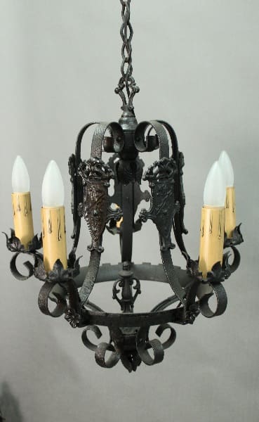 A 14408. Spanish Revival Chandelier With Shield Motif with six candle-shaped bulbs, featuring ornate metalwork and hanging from a chain against a gray backdrop.