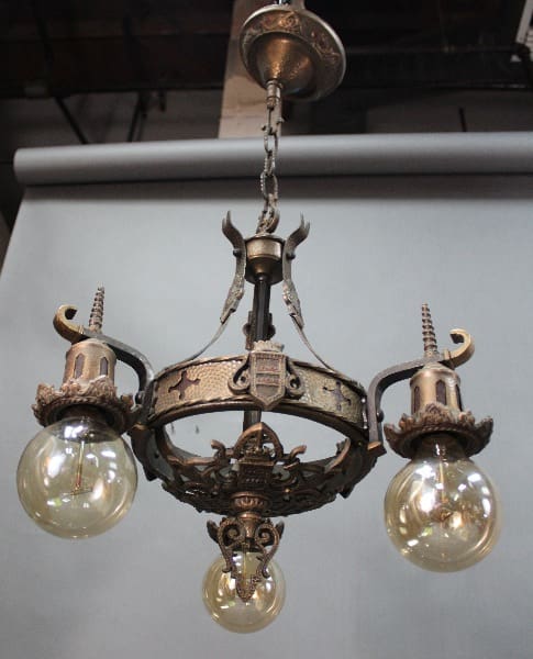 14530. 1920's Three Light Chandelier With Crest Motif with three ornate metallic arms and round clear light bulbs, suspended from a ceiling by a chain.