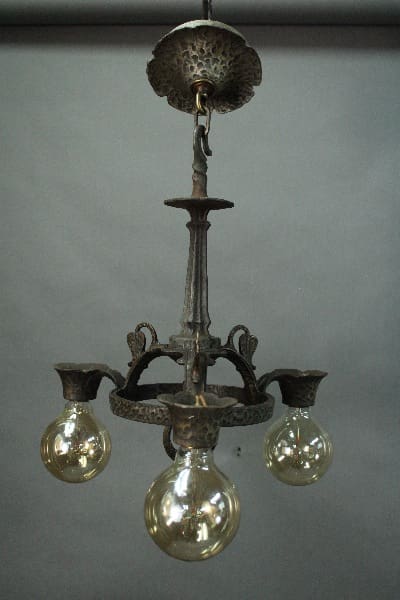 14601. Spanish Revival Three Light Chandelier with three lit bulbs, ornate metalwork, hanging from a ceiling mount in a gray room.