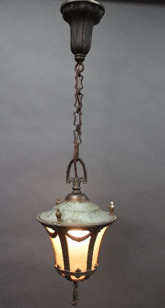 Turn Of The Century Pendant With Original Glass hanging lantern with a lit bulb, suspended by a chain from a dark ceiling mount, against a grey background.