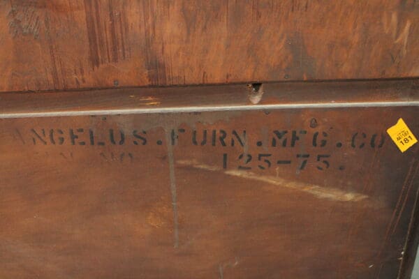 Close-up of a wooden furniture piece with "14696. Monterey Period Angelos Furniture Company Hutch" stamped on it, featuring a yellow price tag.