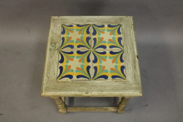 A wooden stool with a distressed finish, featuring a square, colorful tile with a blue and orange floral design on the seat. 15250. 1920's California Tile Table With Four Geometric Tiles.