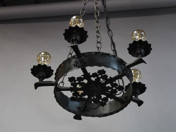 A rustic chandelier made of black metal, featuring floral designs and five exposed light bulbs, hanging from a ceiling by chains. This is the 15308. 1920's Five Light Chandelier.