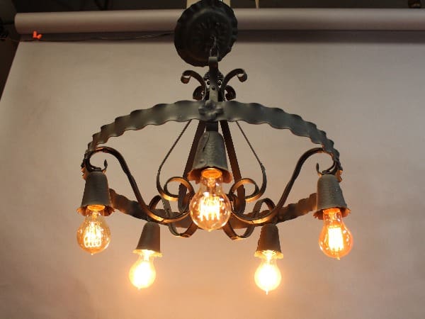 15370. 1920's Spanish Revival Chandelier with five glowing edison bulbs, mounted on a ceiling with a plain backdrop.