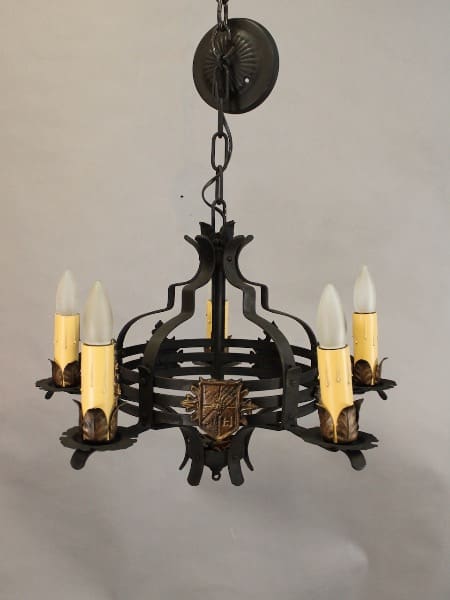 A vintage Spanish Revival chandelier with five candle-shaped lights, featuring a central heraldic shield motif and ornamental details.