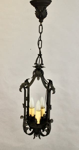 An ornate hanging chandelier with a dark metal frame, featuring gothic-style arches and three candle-like bulbs, suspended from a chain against a grey background - 15497. 1920's Three Light Pendant With Acanthus Leafs.