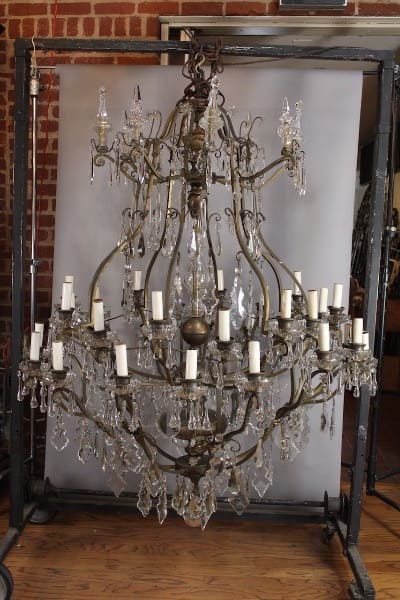 Antique 15605. Monumental Large Scale Crystal Chandelier Circa 1920's 4 FT wide with intricate metalwork and crystal decorations, displayed against a gray backdrop in a room with brick walls.