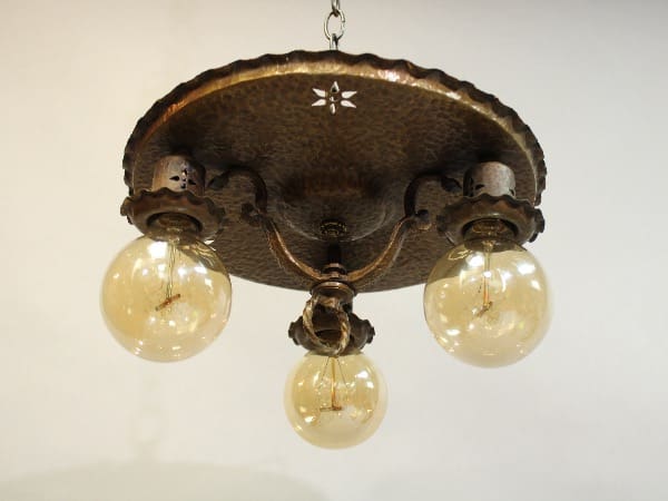 Antique 15949. Hammered Brass Three Light Ceiling Mount with three glowing bulbs, ornate metal detailing, and a central decorative finial, hanging against a plain background.