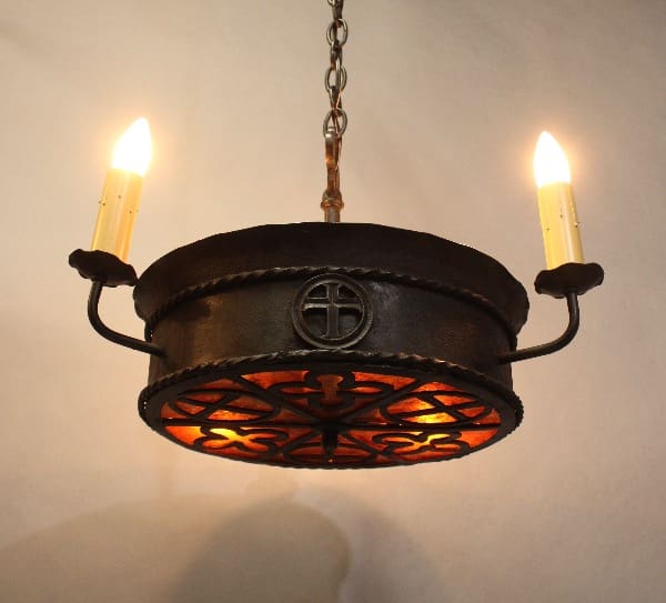 A rustic chandelier with a circular iron frame, featuring the 16250. 1 of 2 Spanish Revival Pendant With Mica emblem, two lit electric candles, and ornate cut-outs casting soft shadows.