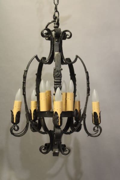 A 16310. Wrought Iron 1920's Spanish Revival Chandelier With Acanthus Motif with six candle-shaped light bulbs, suspended by a chain against a gray backdrop.