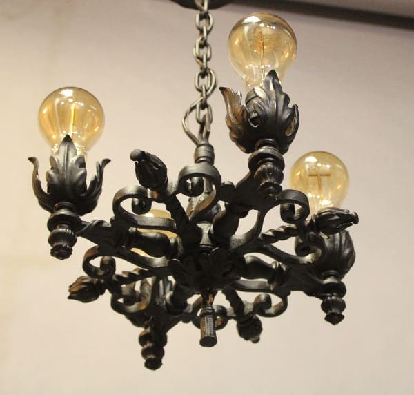 An ornate black iron 16416. 1920's Small Chandelier With Acanthus Motif with three illuminated bulbs, suspended from a chain against a light background.
