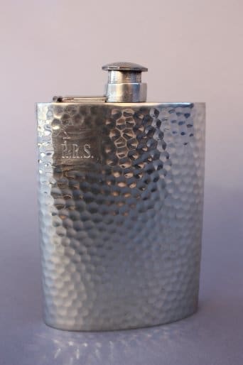 A 7437. English Pewter Flask with a hammered texture and initials "j.r.s." engraved on the front, set against a light purple background.