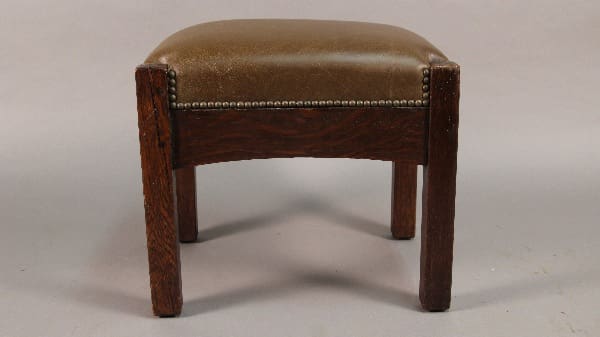 A small, vintage Antique Mission Arts And Crafts Arched Footstool with a cushioned, gold-toned seat and decorative metal studs along the edges.