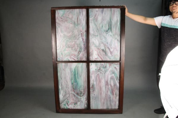 A person in a studio presenting a large, vertical 11324. 1 Of 2 Large Turn Of The Century Four Panel Stained Glass framed in dark wood.