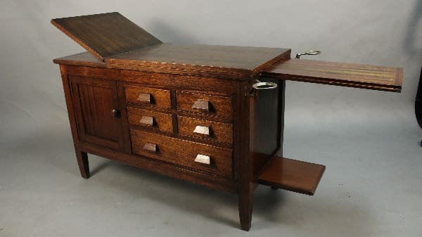 11436. Circa 1910 Arts And Crafts Oak Medical Examination Table with an open lid and pull-out writing surface, featuring multiple drawers and brass handles.