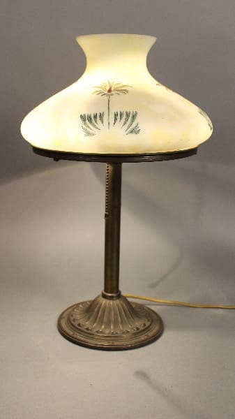 Vintage 11751. Arts And Crafts table lamp with a floral design on a creamy shade, featuring a brass base, illuminated, against a grey backdrop.