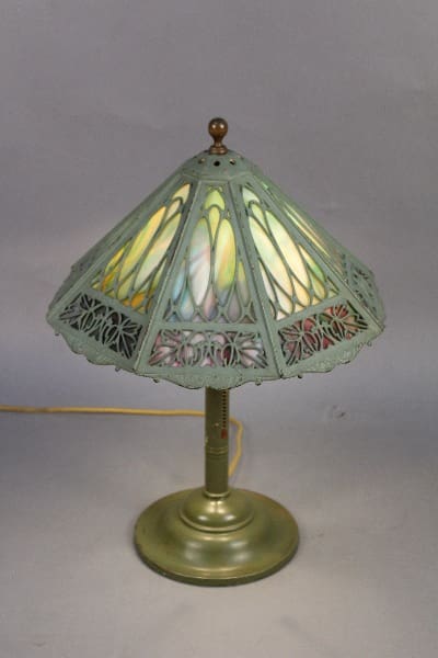 11754. Bradley and Hubbard Signed stained glass table lamp with a brass base and a greenish shade featuring intricate cut-out designs, displayed against a gray background.