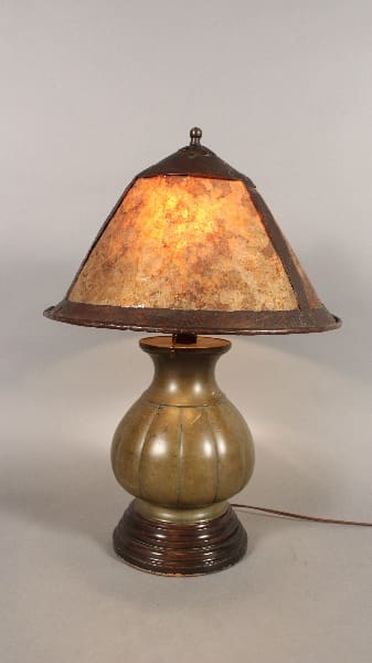 An arts and crafts table lamp with bronze Japanese base and mica shade, illuminated, on a neutral background.