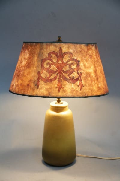 A table lamp with a yellow base and an aged paper shade featuring a red and brown floral design, illuminated in a dimly lit room. 
Product Name: 12021. School of North Dakota Lamp With Beautiful Period Mica Shade
