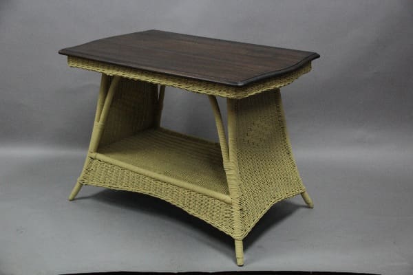 A small, angled Turn Of The Century Wicker Table with a lower shelf, set against a gray backdrop.