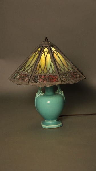 A 12226. Turn Of the Century Art Pottery Lamp with Slag Glass Shade and a turquoise ceramic base, illuminated and set against a dark background.