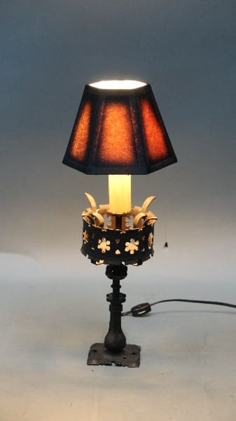 12268. Spanish Revival Wrought Iron Table Lamp with a lit bulb, featuring a dark fabric shade and ornate metal base with floral accents, set against a light gray background.