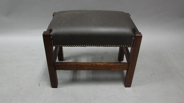 A small, Arts And Crafts 1910 footstool with new leather upholstery and brass stud detailing, positioned centrally against a grey background.