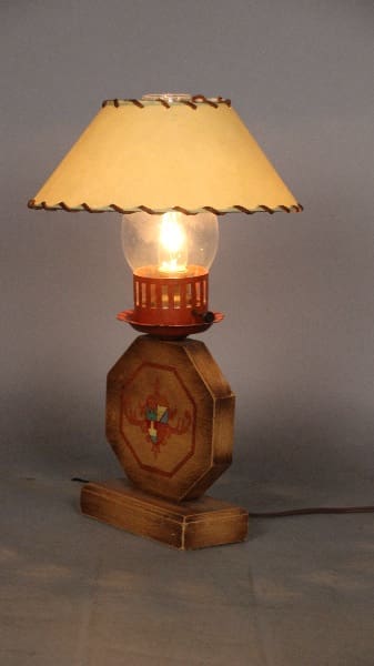 An illuminated 12661. Monterey Period Table Lamp with an octagonal wooden base featuring an inlaid red cross design, supporting a cream-colored shade.
