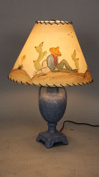 12673. 1920's Table Lamp With Hand Painted Shade with a blue base and a shade depicting a cowboy sitting under a cactus, illuminated from within, set against a gray background.