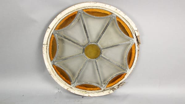 Round stained glass window with a star-shaped design in white and amber, showing signs of age and wear, against a plain background.