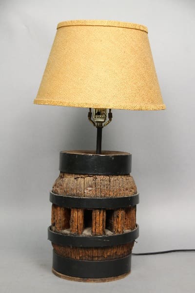 13498. Rustic Table Lamp Circa 1920's with a shade on top of a rustic, barrel-like wooden base, placed against a gray backdrop.