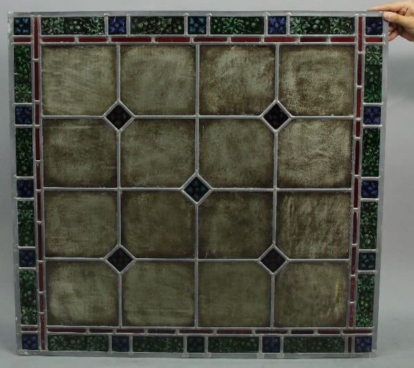 A 13691 stained glass window featuring a geometric pattern with hexagonal shapes, bordered by red framework and small colorful glass accents, held by a hand on the right side.