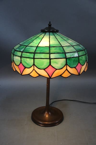 A 14011. Stained Glass Shade Table Lamp with a bronze base Circa 1910, featuring green and pink sections, illuminated and standing against a dark background.