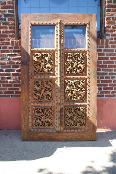 Sentence with Product Name: Ornate wooden double doors with intricate carvings, standing against a brick wall, 14490. Monumental Carved Door With Glass Salvaged From Beverly Hills Home.