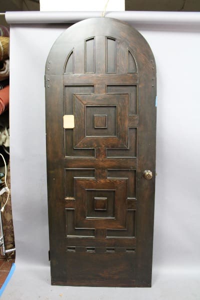 Sentence with product name: A 14765. Spanish Revival Arched Door #7 with geometric patterns and a brass knob, standing against a cluttered background.