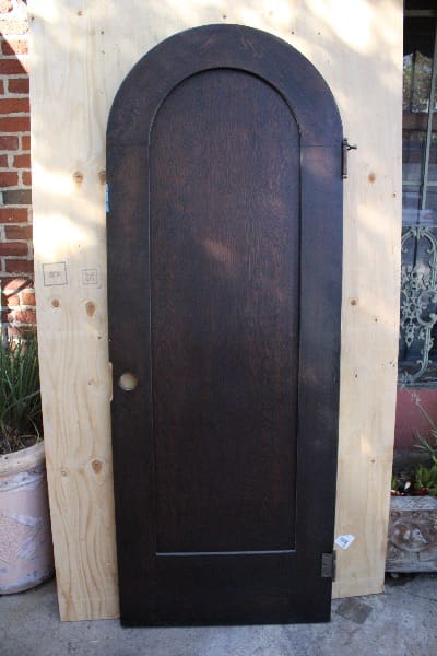 A tall, dark 14772. Spanish Revival Arched Door # 6 propped against a plywood board outside, with a metal hinge visible on one side.