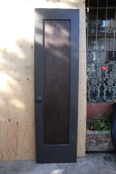 A dark brown 14773. Narrow Spanish Revival Door #11 with a vertical window, set in a plywood-covered wall, adjacent to decorative metal grilles and foliage.