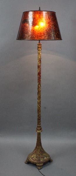 An ornate 14803. 1920's floor lamp with a carved wood base with gilt finish and a glowing amber lampshade, standing against a neutral gray background.