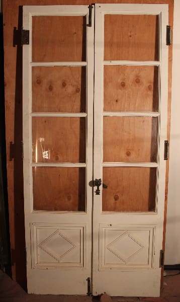 A pair of vintage white wooden doors with glass panes on top and solid panels on the bottom, set against a simple plywood backdrop. 14818. 1 of 6 Set of French Doors Circa 1920's With Diamond Motif