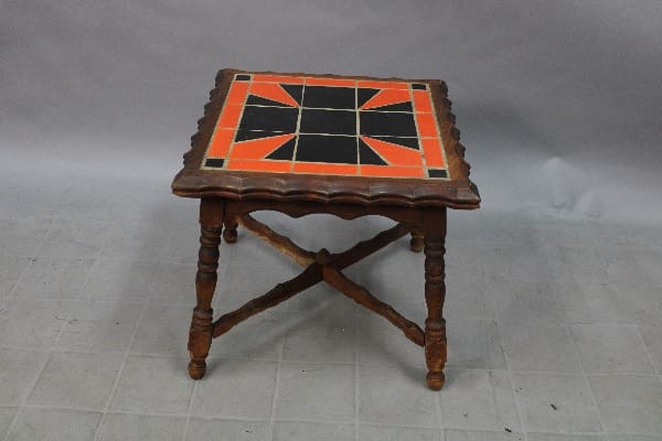 Small wooden side table with a black and orange 14926. 1920's California Tile Table top, featuring crossed leg design, set against a gray backdrop.