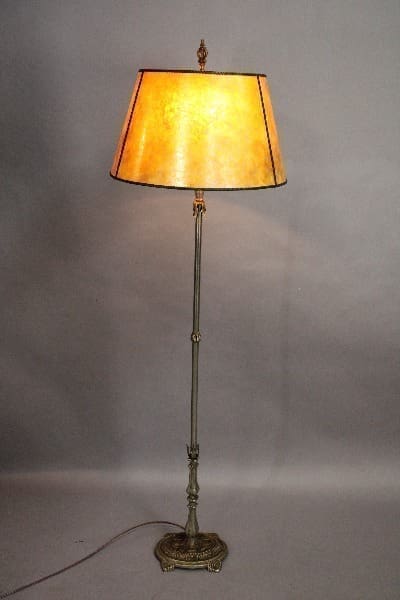 A tall, antique floor lamp with an illuminated orange shade, ornate brass base, and a visible power cord against a neutral background.