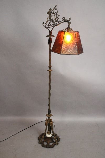 Antique Spanish Revival Bridge lamp with ornamental metal design and a red stained-glass shade, lit and standing on a gray background.